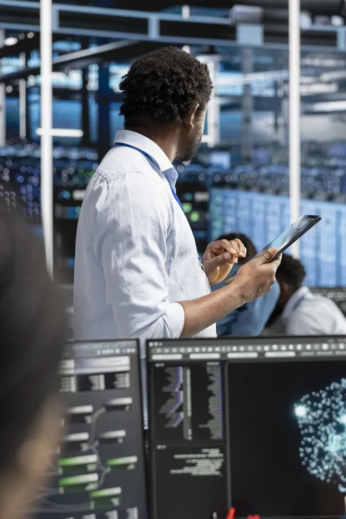 IT administrator using a tablet to oversee AI-powered system operations in a modern data center surrounded by digital monitoring screens.
