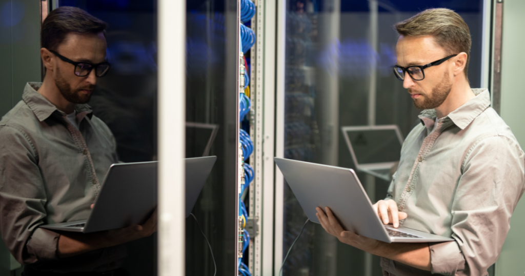 man standing with a laptop next to servers at The Nu-Age Group.